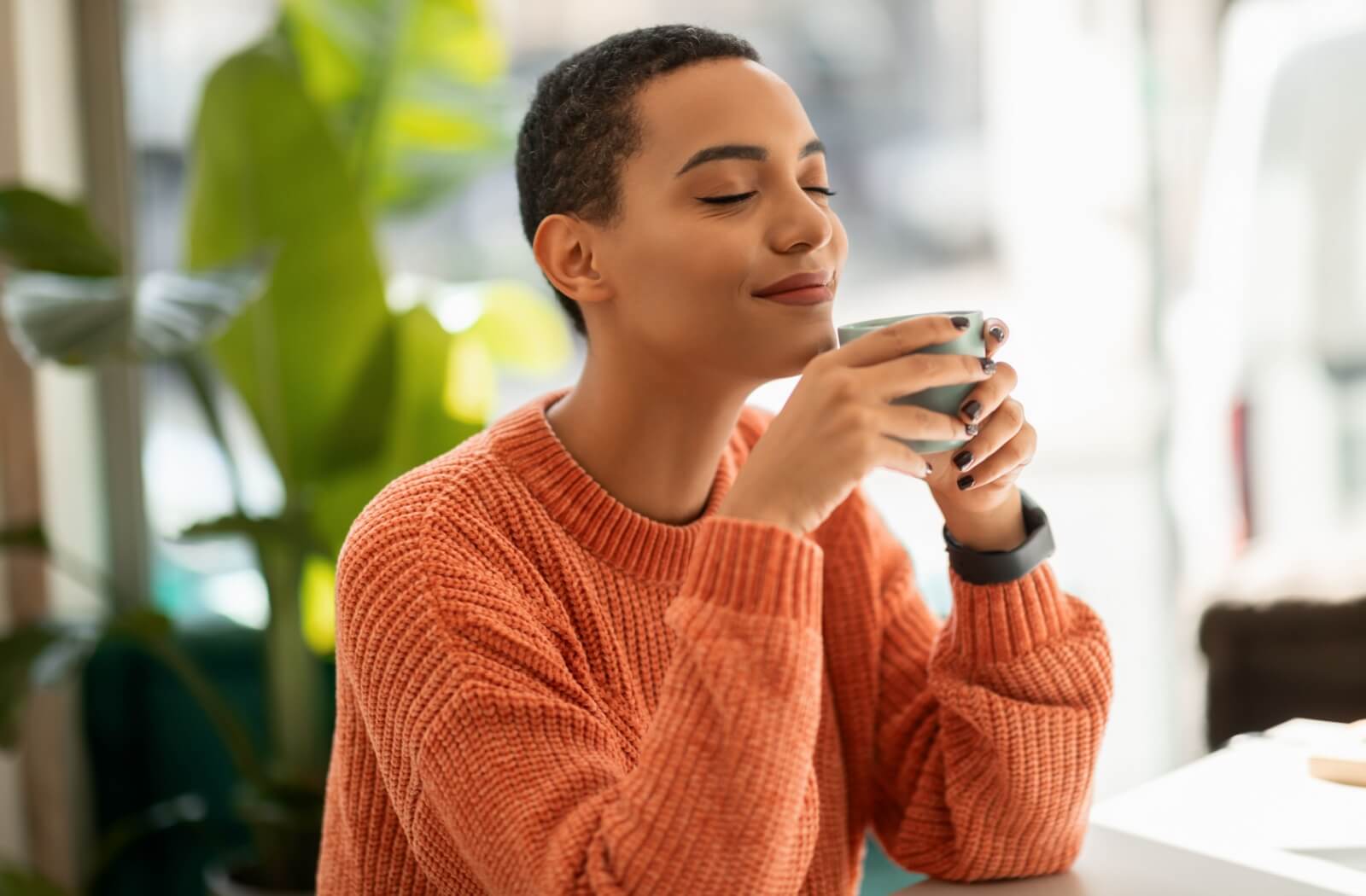 Relaxed young adult holding cup with both hands to their face while smiling with eyes closed.