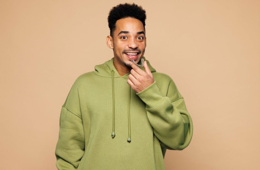 Smiling young adult in green hoodie holding clear aligners (Invisalign) to mouth against a pale orange background.