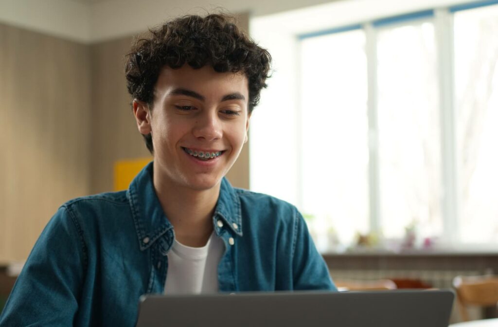 Person with curly hair and metal braces smiling while looking at a laptop in a bright indoor setting.